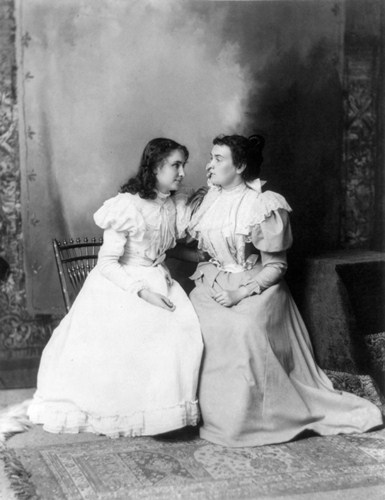 foto antiga em preto e branco de Hellen Keller sentada numa cadeira com uma das mo no rosto de Anne Sullivan